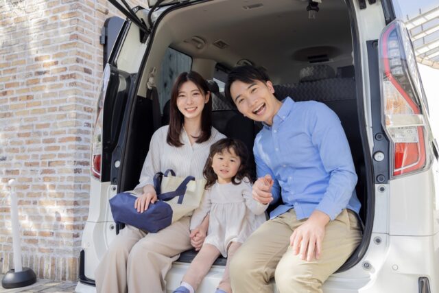 久米島でのレンタカー旅行を楽しむ笑顔の家族。詳細白い車のバックドアを開け、荷室の縁に座るお父さん、お母さん、小さな女の子。家族旅行の合間のリラックスした雰囲気。
