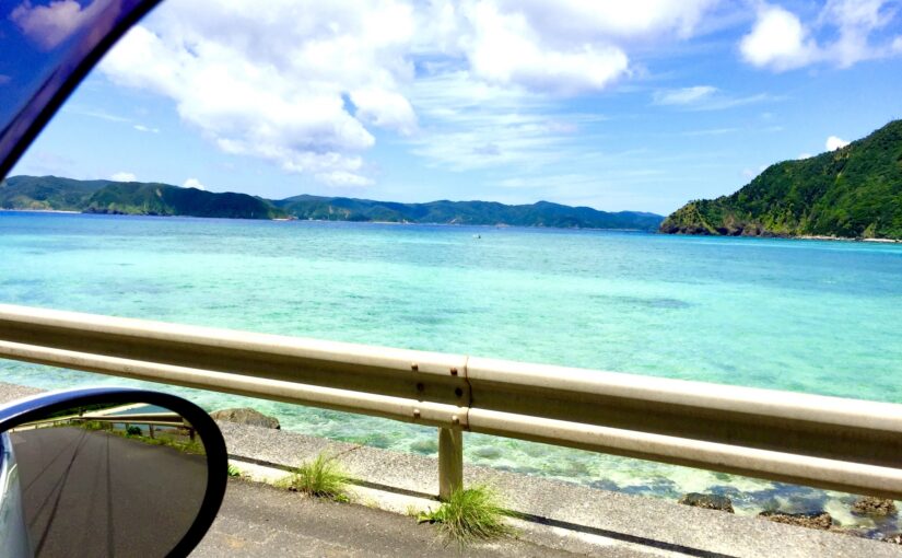 レンタカーの車窓から見える久米島の透き通ったエメラルドグリーンの海と緑の山々