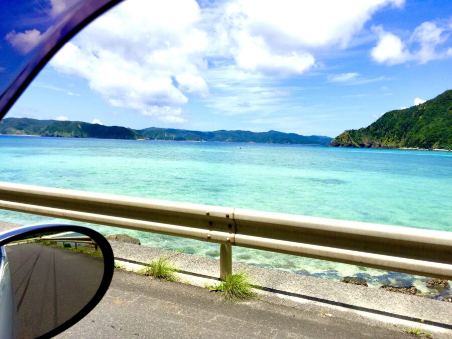 レンタカーの車窓から見える久米島の透き通ったエメラルドグリーンの海と緑の山々